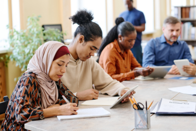 In an office, we see a group of 4 people sat at grey rectangular table, taking notes on notepads, and one man standing behind them, looking at his phone.