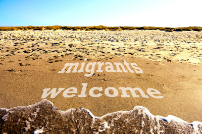 Text imposed over the wet sand on a beach, the incoming wave almost touching it. In white lowercase, it reads "migrants welcome," On the horizon is a line of brown rocks, in front of a clear blue sky.