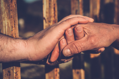 Two people clasp hands, separated by a rusted barred fence.