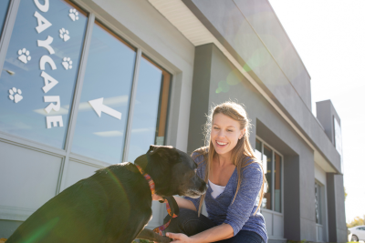 Smiling woman and her dog stand outside a building with the word "daycare" printed on a window.  