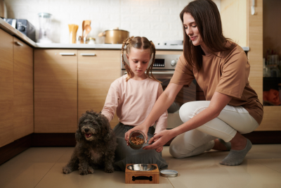 A mother, daughter, and grey doodle puppy sit on the floor of a kitchen. The Woman, with long brown hair, holds a scoop of dog food in her hand. She's tipping the kibble into a metal bowl. Her daughter, who has braided pigtails in her ash blond hair, watches intently. The little girl also rests her hand on the pups back, while the puppy sits beside her, leaning into her.