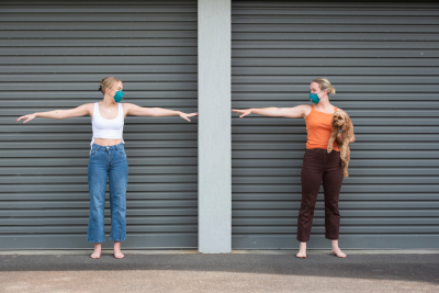 Two white women wearing cyan blue masks, with long blonde hair swept into low buns, socially distancing.  The woman on the left extends both arms out to her sides, and looks to the woman on the right.  The rightmost woman reaches out the hand closest to the other woman, and cradles a light brown doodle in her other hand.  