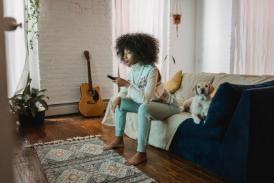 Sitting on a couch, a light-skinned black woman with mid length chestnut curls in an afro blowout holds a TV Remote. Next to her a yellow lab puppy lays, looking off into the distance. The woman leans forward, feet planted on the ground, with a focused expression on her face.