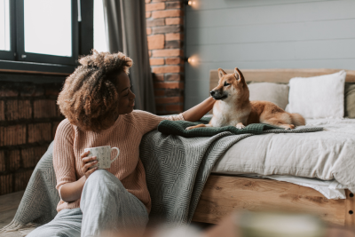 A woman pets her Shiba Inu laying on her bed, while she sits in a chair, balancing a mug on her knee.