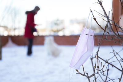 In the center of an exterior shot, out of focus, a heavily bundled up human holds out a hand to a fluffy white dog sitting in front of them. To the left, in focus, bare shrub branches lean into view. A pink disposable face mask, hooked over a twig blows in the breeze.