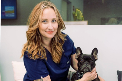 Dr. Knox, a white woman wearing dark blue scrubs with long golden wavy hair, softly smiles while holding a black french bulldog, who stares ahead calmly. The pair sits in an vet office waiting room.