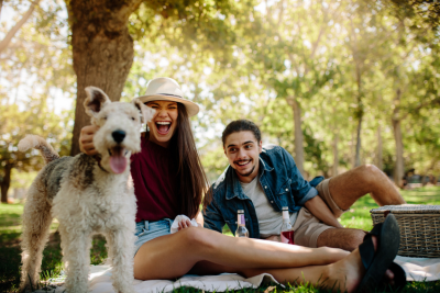 male and female sitting on picknic blanket with their dog smiling. trees and greenery in the background