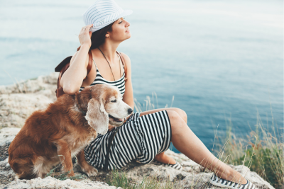 Senior dog with floppy ears sits on rocks with a woman.  They admire the body of water behind them.